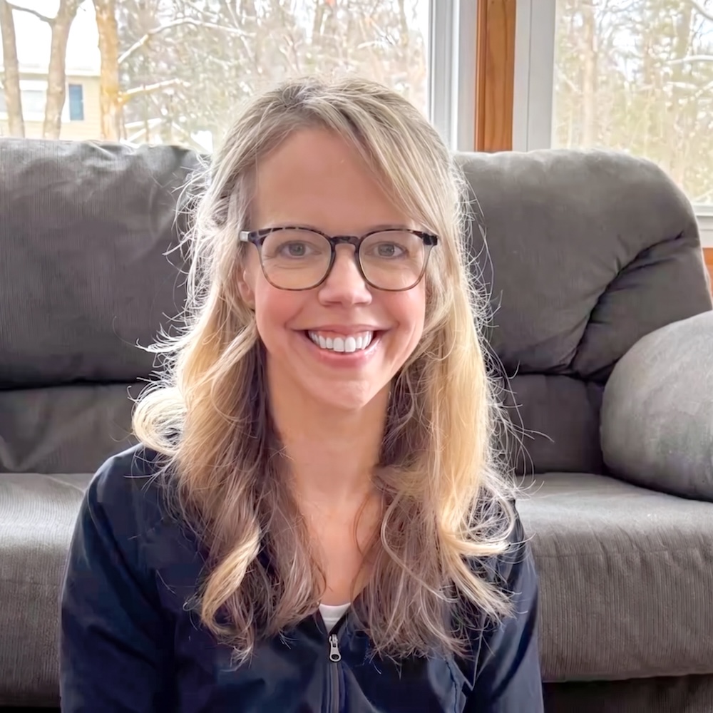 A woman with long blond hair and glasses sits in front of a gray couch, smiling at the camera. Trees and snow are visible through the window behind her.